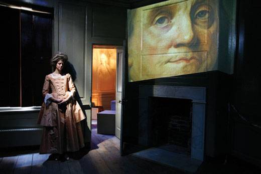 Polly, in a Georgian silk dress, beside projection on the wall