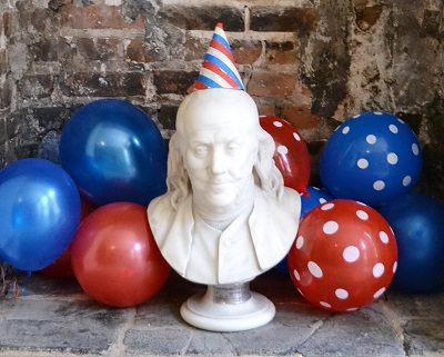 Benjamin Franklin bust with party hat on and red and blue balloons